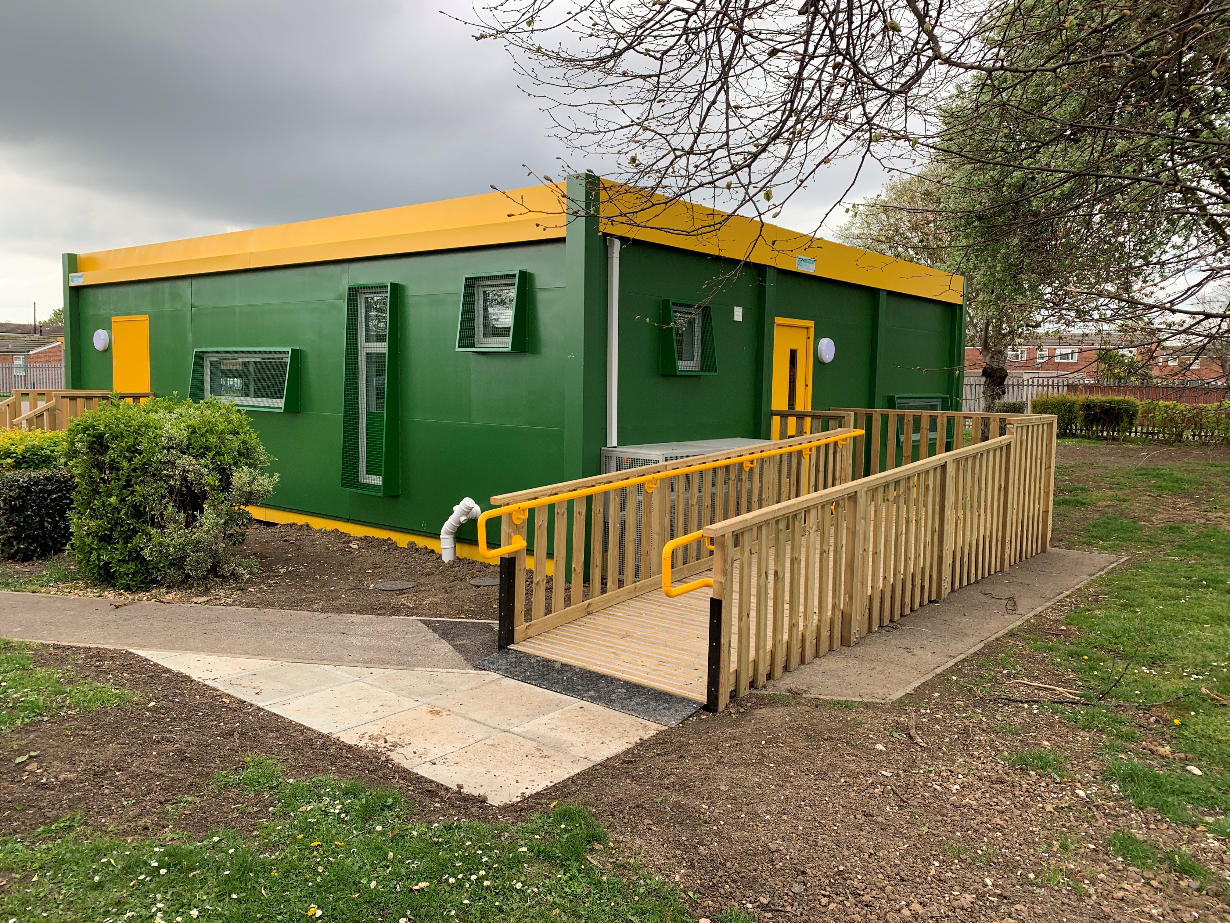 Green and yellow modular classroom with wooden ramp leading to the doorway.